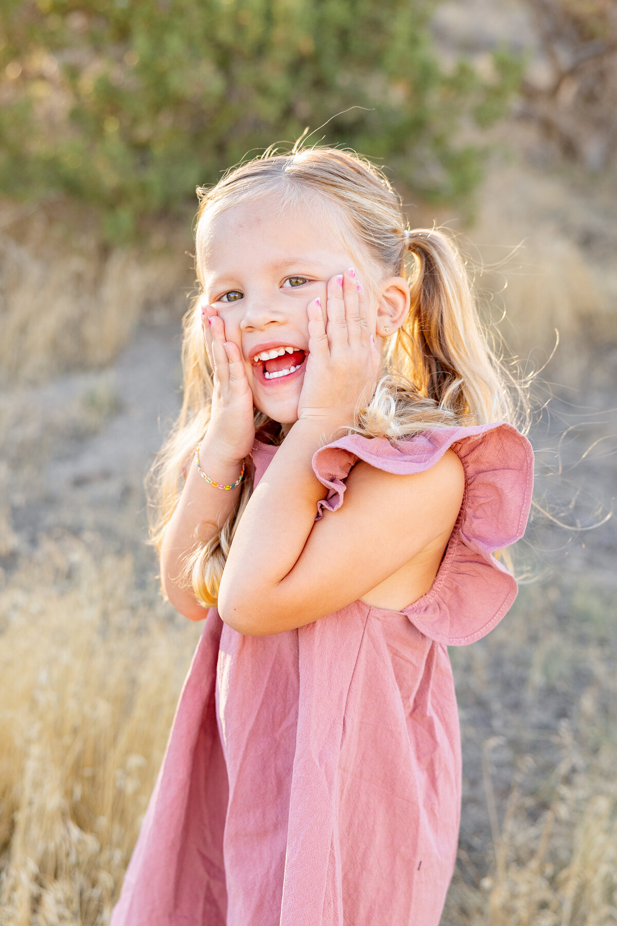A little girl in a pink dress places her hands on her cheeks and laughs, full of excitement and happiness. The golden sunlight and natural background enhance her cheerful expression.