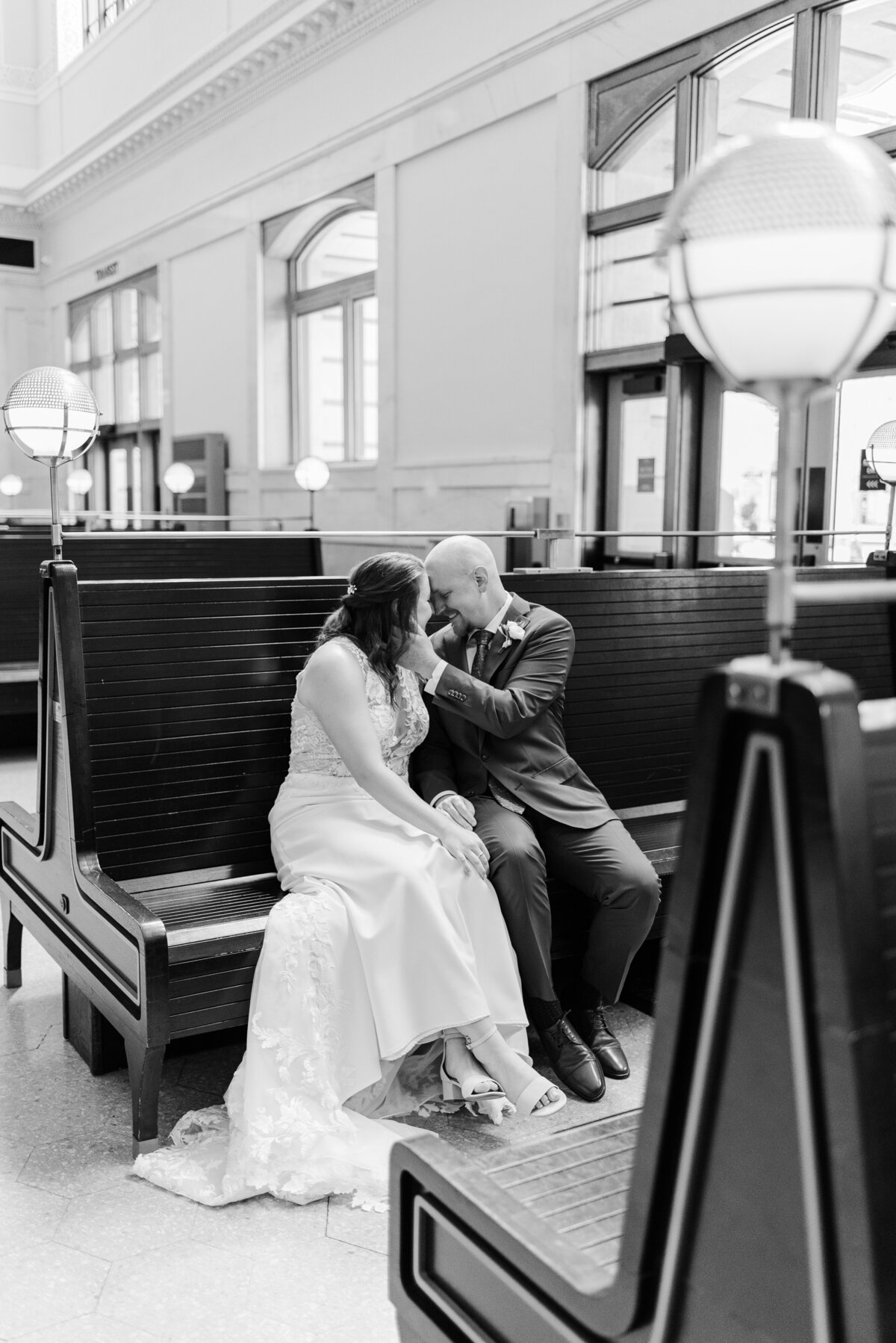 Bride and groom sit on a train station bench while nuzzling close together captured by Denver wedding photographer