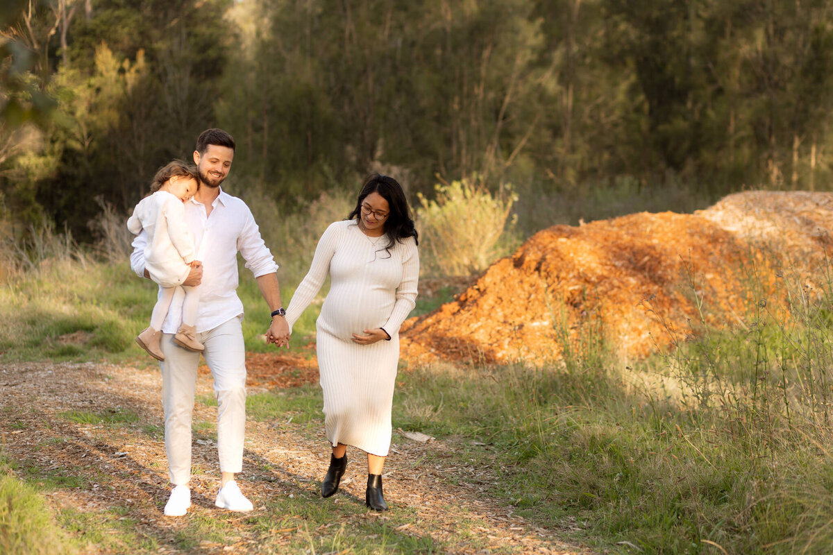 Young family walking through a natural outdoor setting during a maternity photoshoot in Sydney, with father carrying daughter and mother cradling baby bump