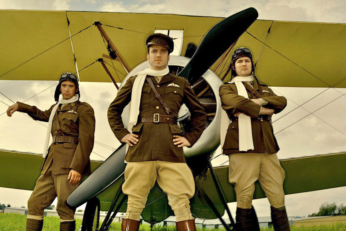Musician portrait trio The Luna Riot wearing vintage pilot outfits standing in front of old propeller plane
