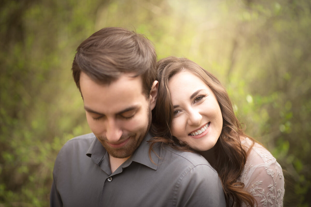 Portrait of an engaged couple standing in a lush green park, with the woman embracing her fiancé from behind.