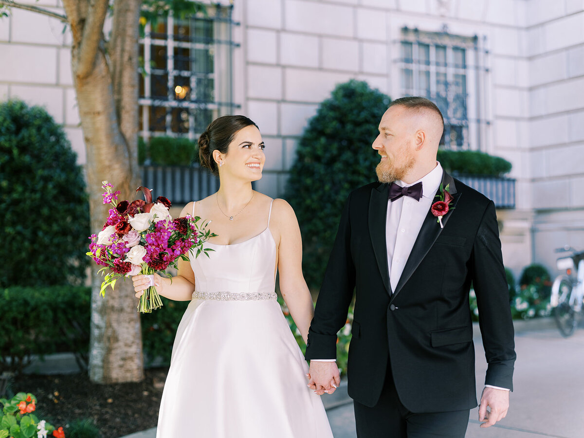 Bride and groom walking hand in hand outside The Hay Adams Hotel DC, with the bride holding a vibrant bouquet of pink, burgundy, and purple flowers, complementing the groom’s classic black tuxedo with a burgundy boutonniere.