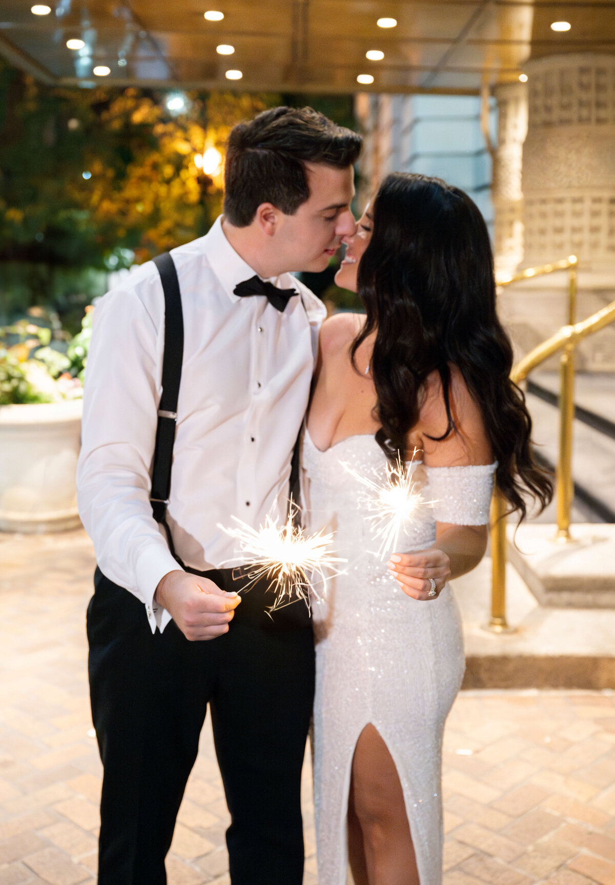 A couple dressed in formal attire shares a romantic moment while holding sparklers. The woman is in a sparkling white gown, and the man is wearing a black bow tie and suspenders. They stand close, with a festive atmosphere around them.
