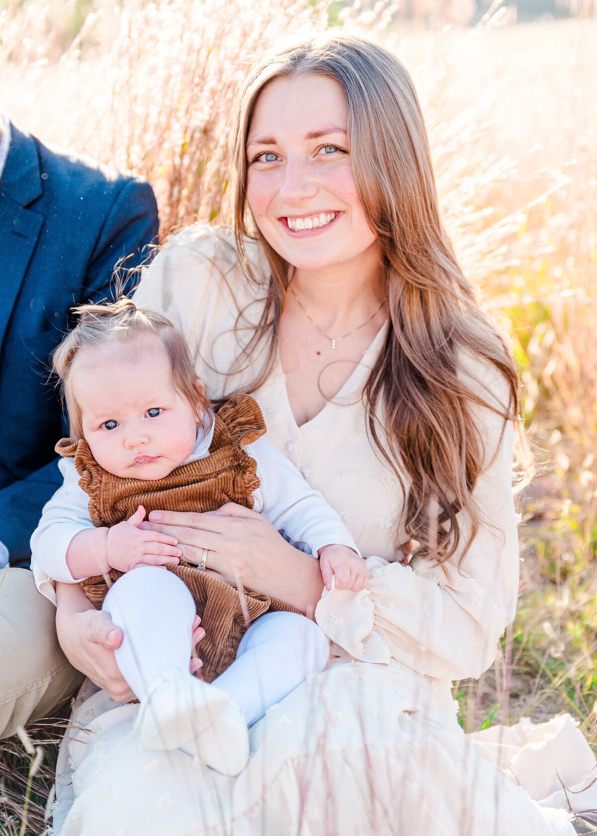 A close up photo of a mother in a tan long sleeve dress smiles up a the camera with her infant daughter in a brown dress  captured by denver family photographer