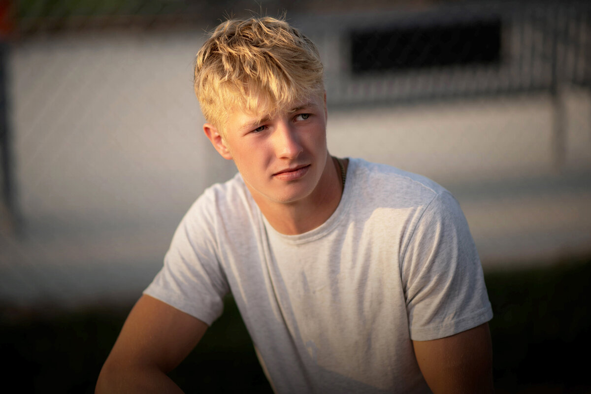 A high school senior in a white shirt sits and leans on his knee in a white shirt in the sun for his Pella Senior Photographer