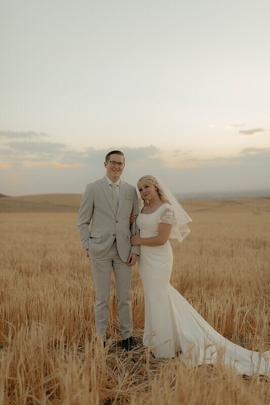 A bride and groom stand next to each other for a portrait. She is holding his arm as they smile at their wedding photographer Brittney Ann Photography.