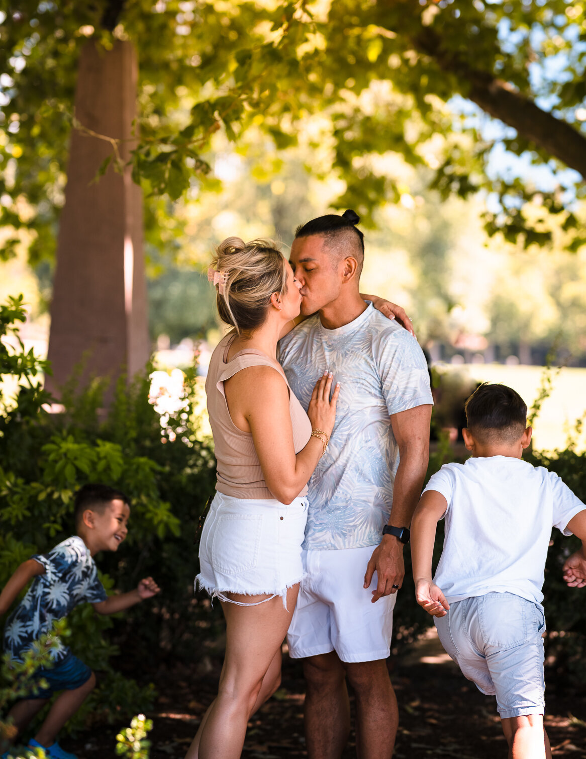 Children having fun running circles around their parents as they enjoy a romantic kiss under a shady tree during their family photoshoot session in downtown Bellevue WA.