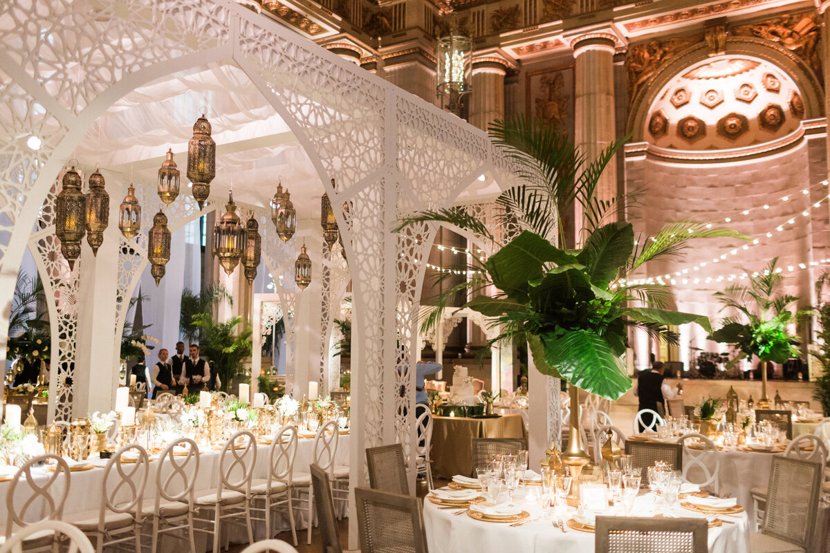 Located in the heart of Washington D.C., The Mellon Auditorium is one of the best luxury venues in the city known for its European flair. The photo captured by photographer Sarah Bradshaw showcases the grandeur of this elegantly decorated wedding reception hall. The ornate white lattice arches, lush green plants, and hanging lanterns add a touch of sophistication to the space.  The round tables are elegantly set with white and gold tableware, complemented by white chairs that add to the luxurious atmosphere. Soft lighting further enhances the opulent ambiance of the venue, creating a captivating setting for a memorable event.