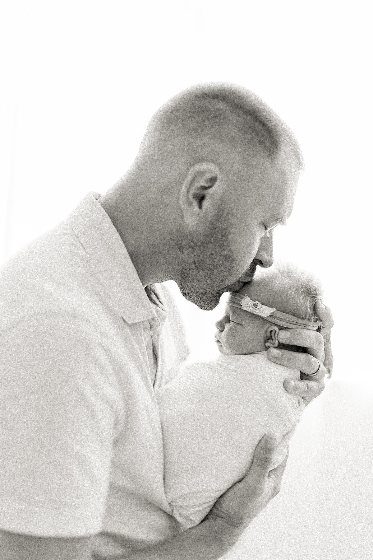 Black and white newborn photo of a father holding his newborn baby girl up as he kisses her head.