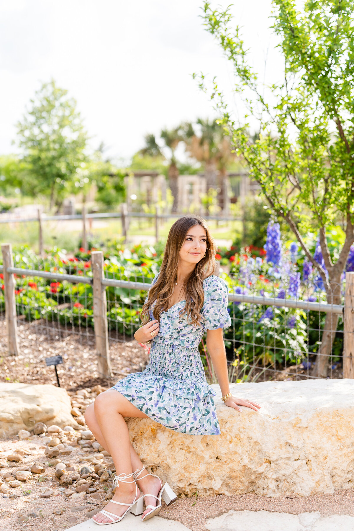 Texas A&M senior girl sitting on stone bench while wearing blue dress and touching hair while looking away at Leach Teaching Gardens