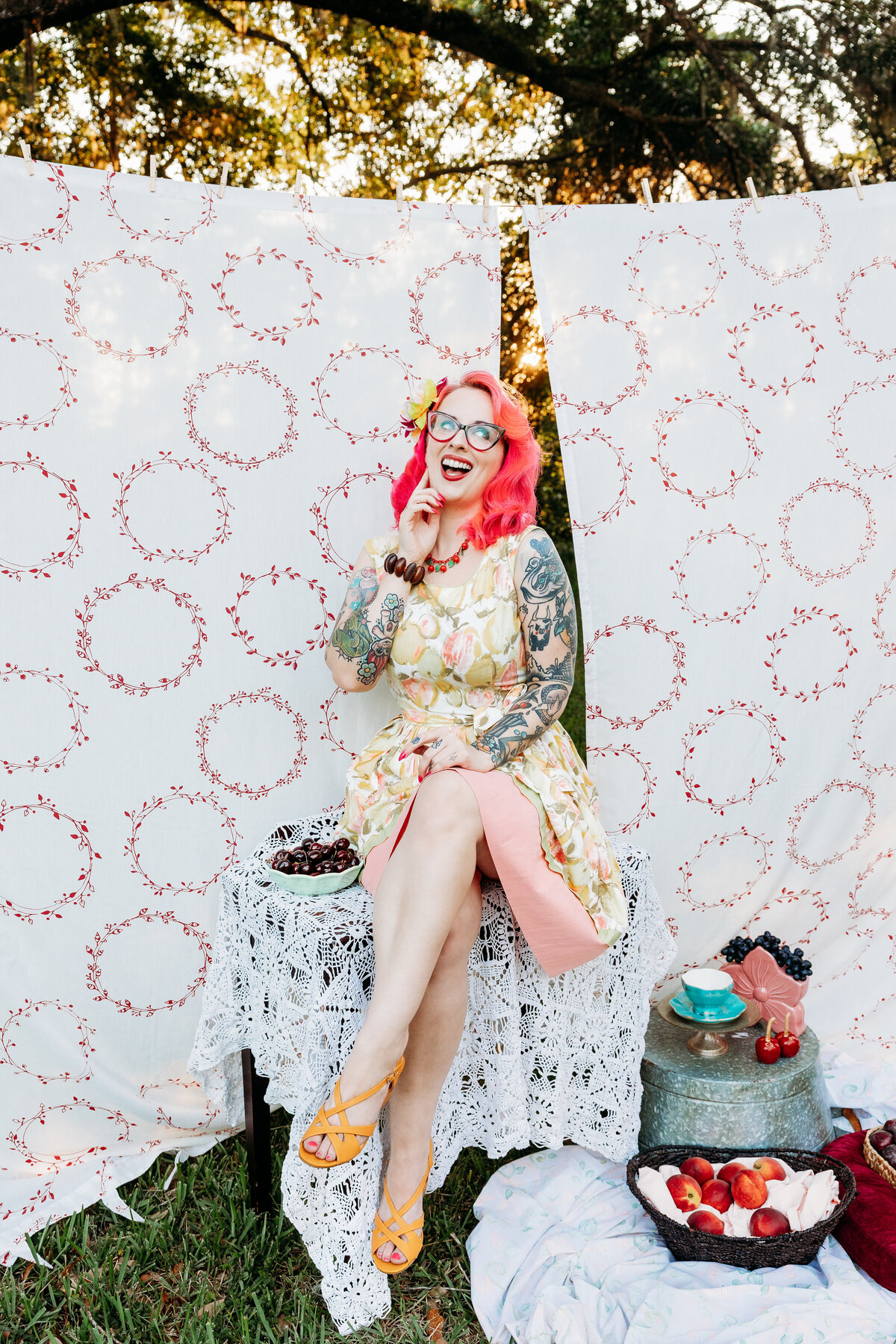 Woman with pink hair and glasses laughs joyfully at a vintage-inspired picnic table with cherries and lace in a Gainesville field.