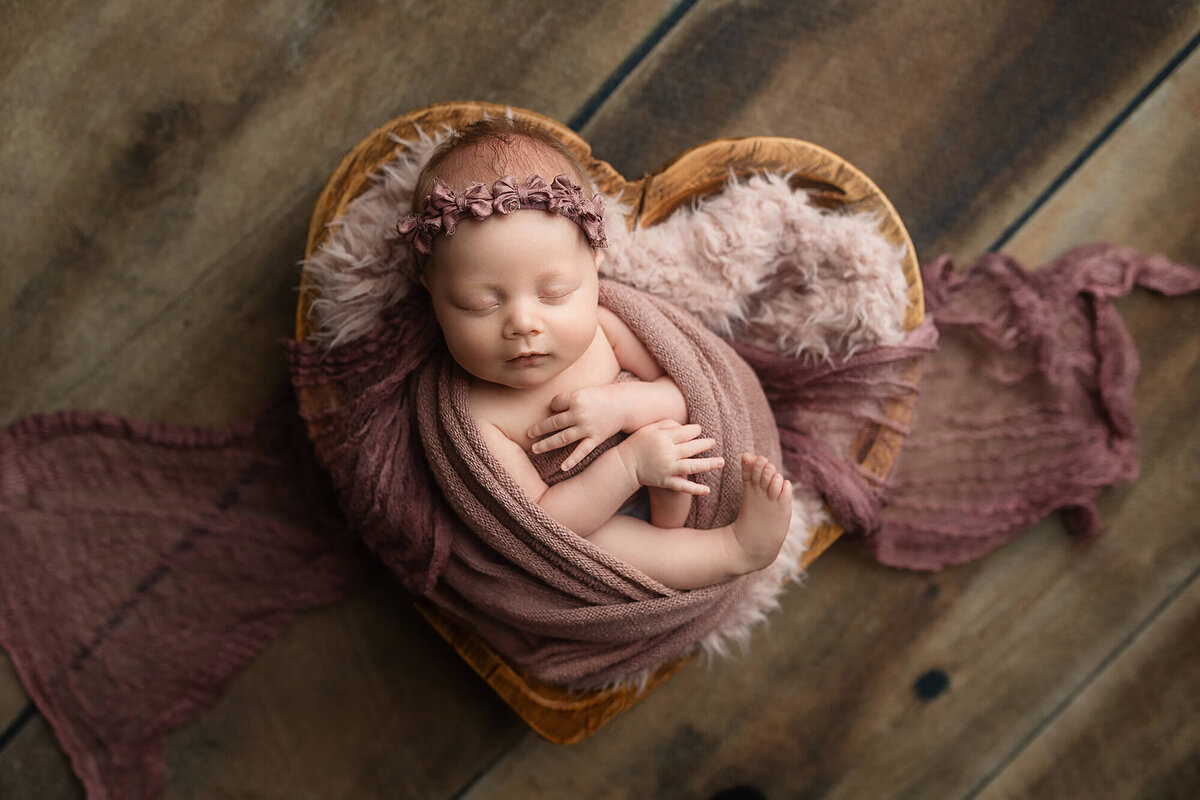 Newborn baby girl wrapped in a lilac color fabric posed in a wooden heart bowl during her Newborn session at Jennifer Brandes Photography.