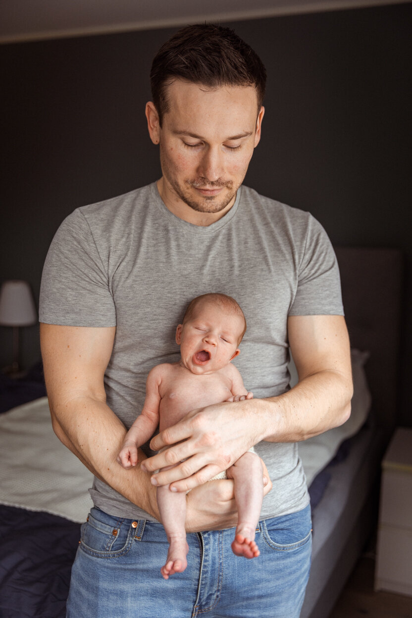 Livsstilsfoto av fars overkropp, han holder babyen sin lent mot brystet sitt, babyen er vendt mot kamera og gjesper.