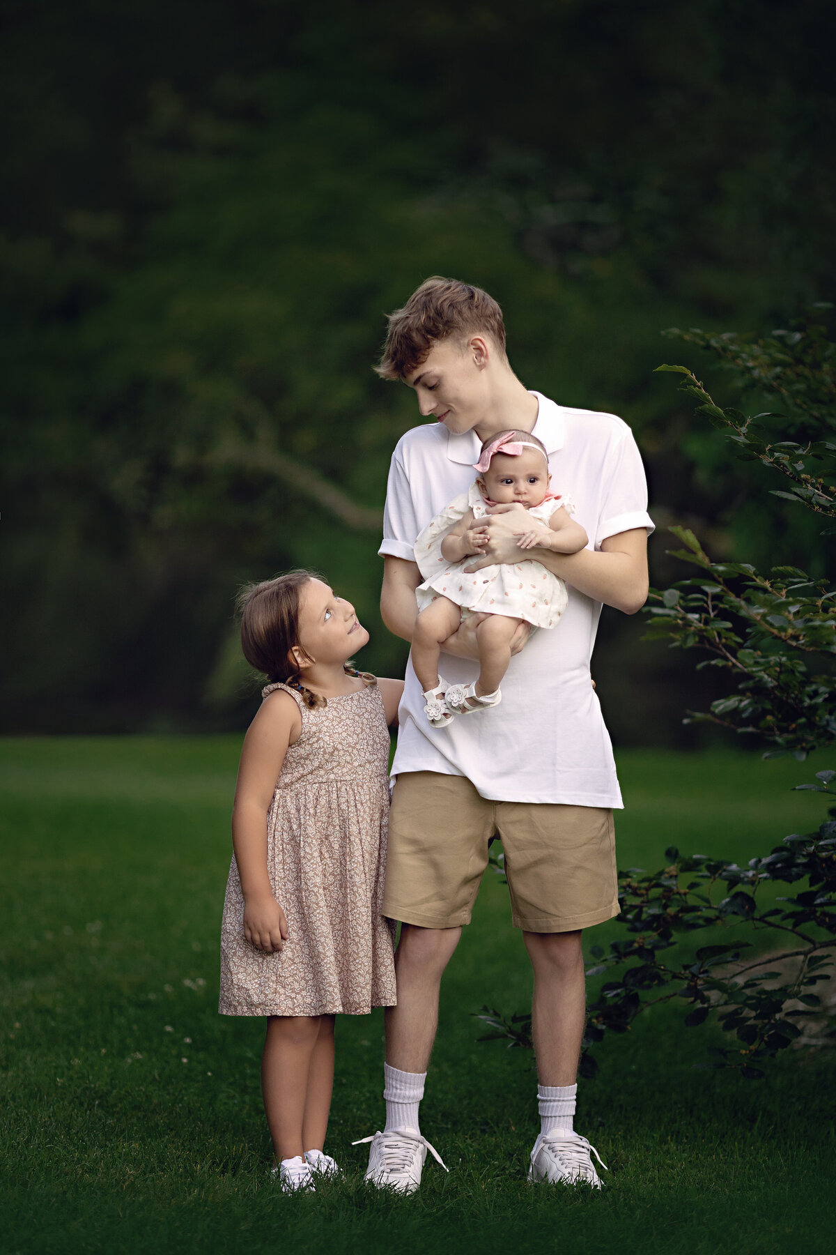 A teen boy in a white shirt stands in a park holding his infant baby sister while his toddler sister hugs onto him