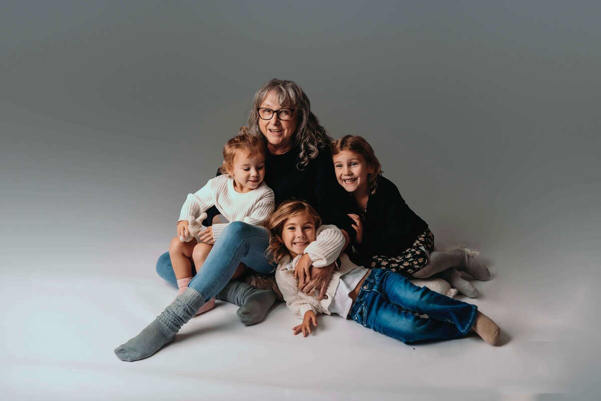 A group photot of a grandmother surrounded by her three granddaughters, aged 2, 4, and 6. They are all wearing cozy sweaters, with the grandmother seated on the floow as the youngest grandchild sits on her lap, the middle grandchild lays beside her with her head resting on her leg, and the oldest grandchild sits beside her, leaning against her shoulder. They are all gathered close, showing their bond and strong connection in this tender moment captured by Mobile, AL Motherhood Photographer.
