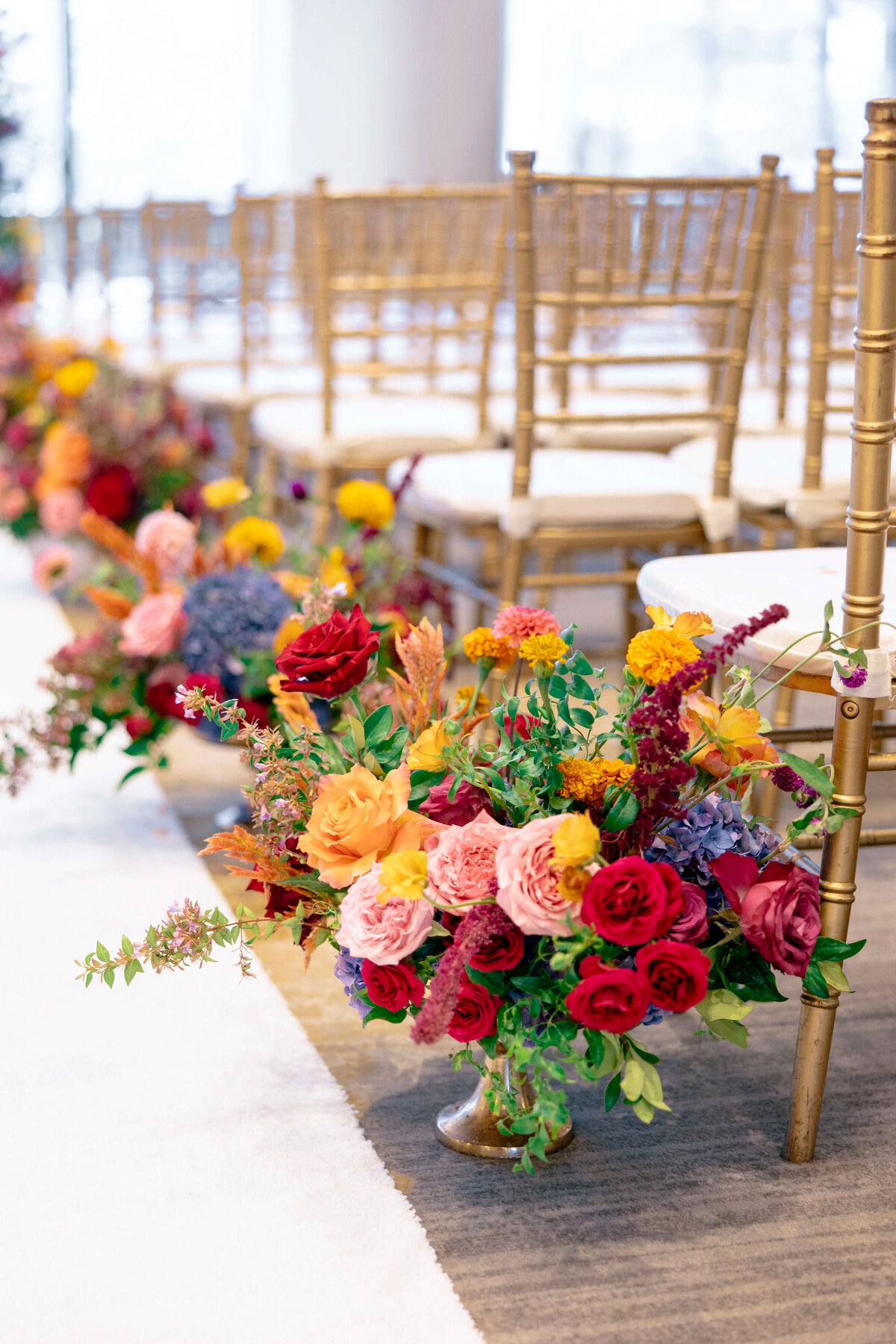 A row of gold chairs lines an aisle adorned with vibrant floral arrangements featuring red, pink, yellow, and orange flowers. Lush greenery adds contrast, creating an elegant setting for a formal event.