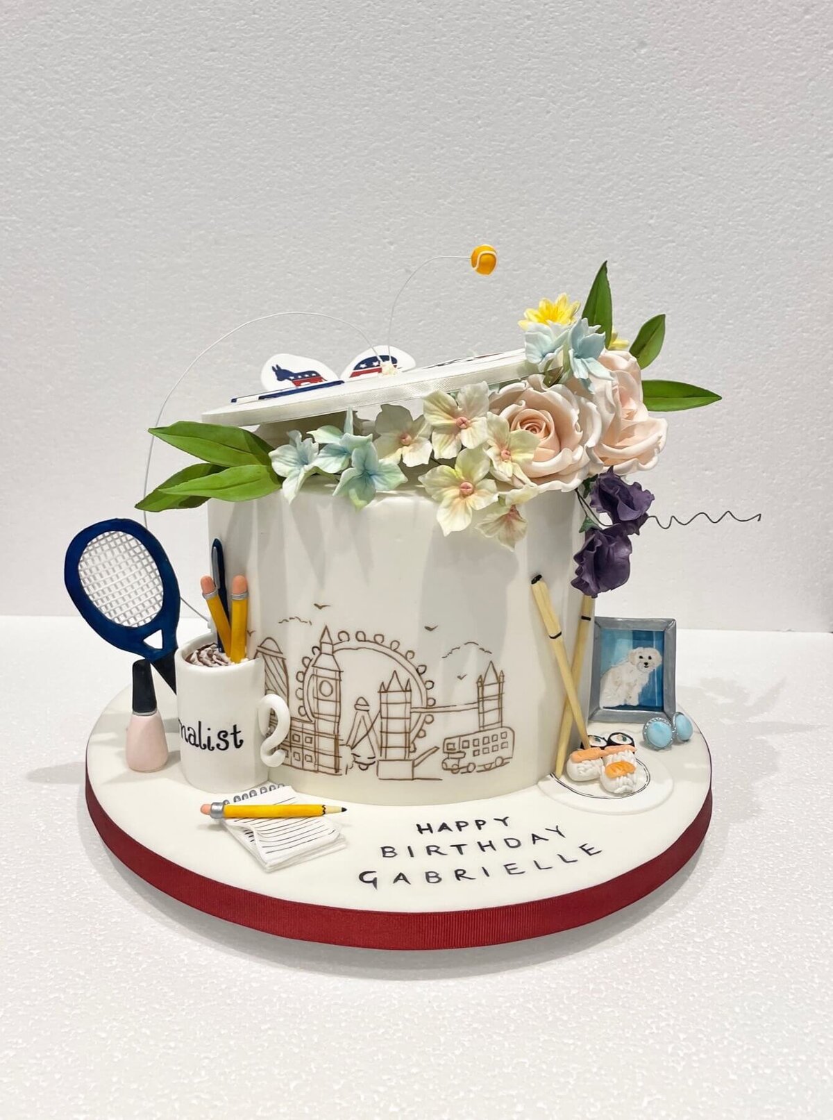 An intricate birthday cake with a hand painted London sky line and a model mug, pencils, tennis racket, nail polish, picture of a dog and more
