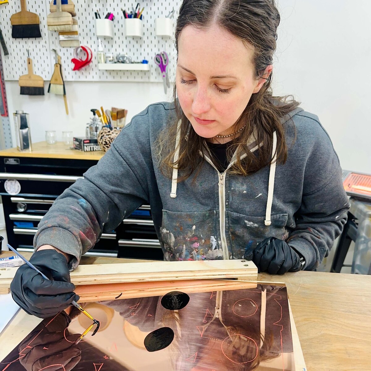 Artist Courtney Sennish in a grey hoodie painting asphaltum on a copper plate with traced shapes, surrounded by printmaking tools and supplies.