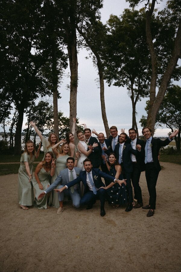 Le couple entouré des garçons et demoiselles d'honneur sur le sable devant un décor de mer et d'arbres. Photo de groupe de mariage par Kamescop.