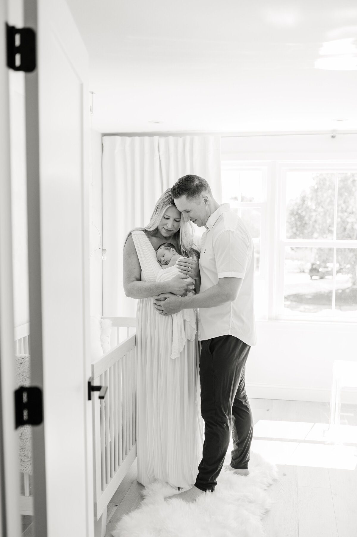This is a pulled back black and white image of mom and dad standing next to their baby's crib while they are holding their baby and looking down at baby. This image is shot through the doorframe.