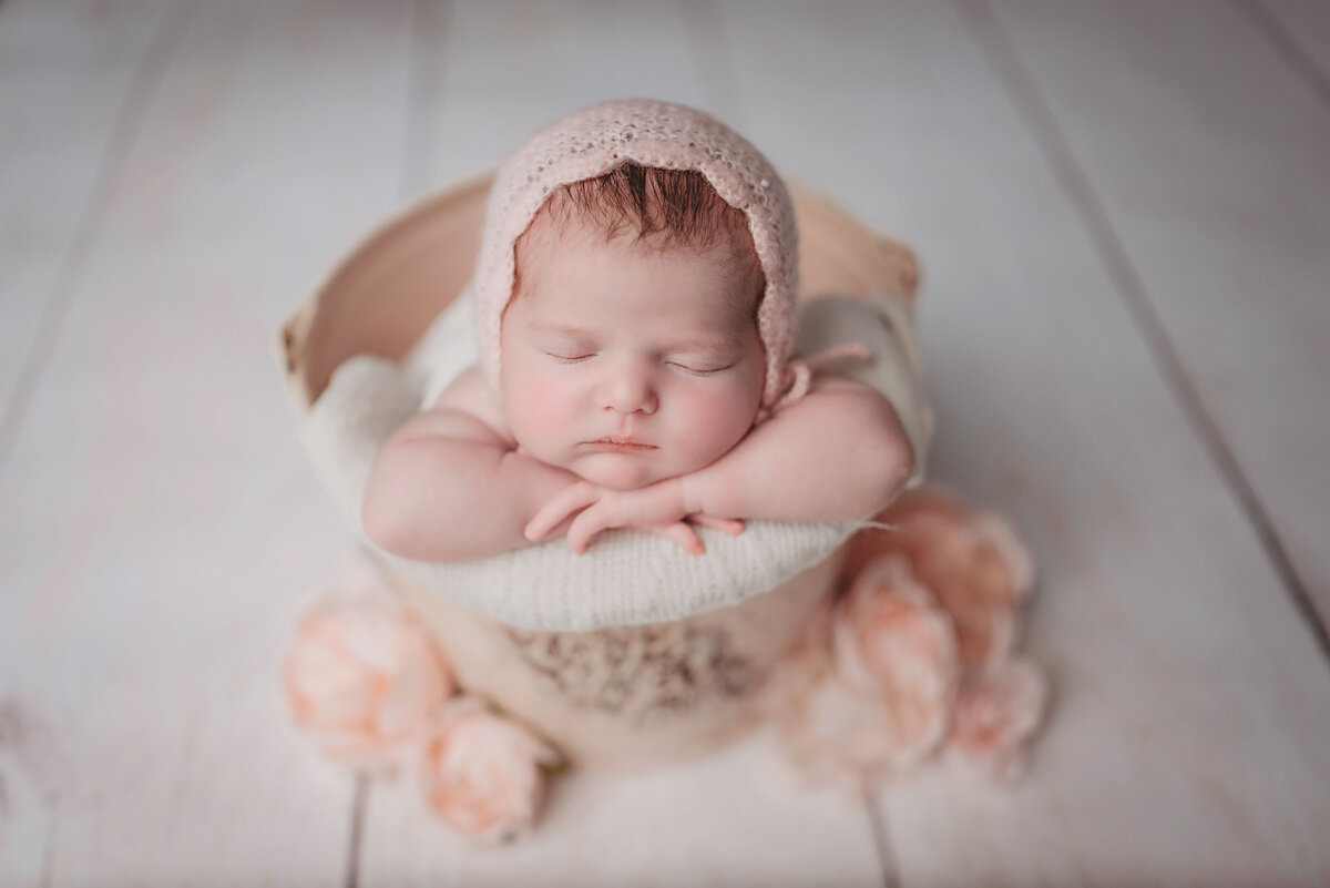 Newborn girl wearing pink scalloped bonnet laying chin on hands in cream color bucket with pink flowers spread around bucket