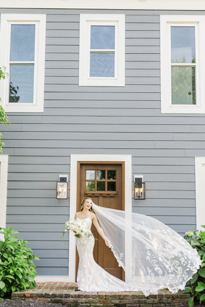 bride smiling in front of a house with her wedding veil flying in the wind