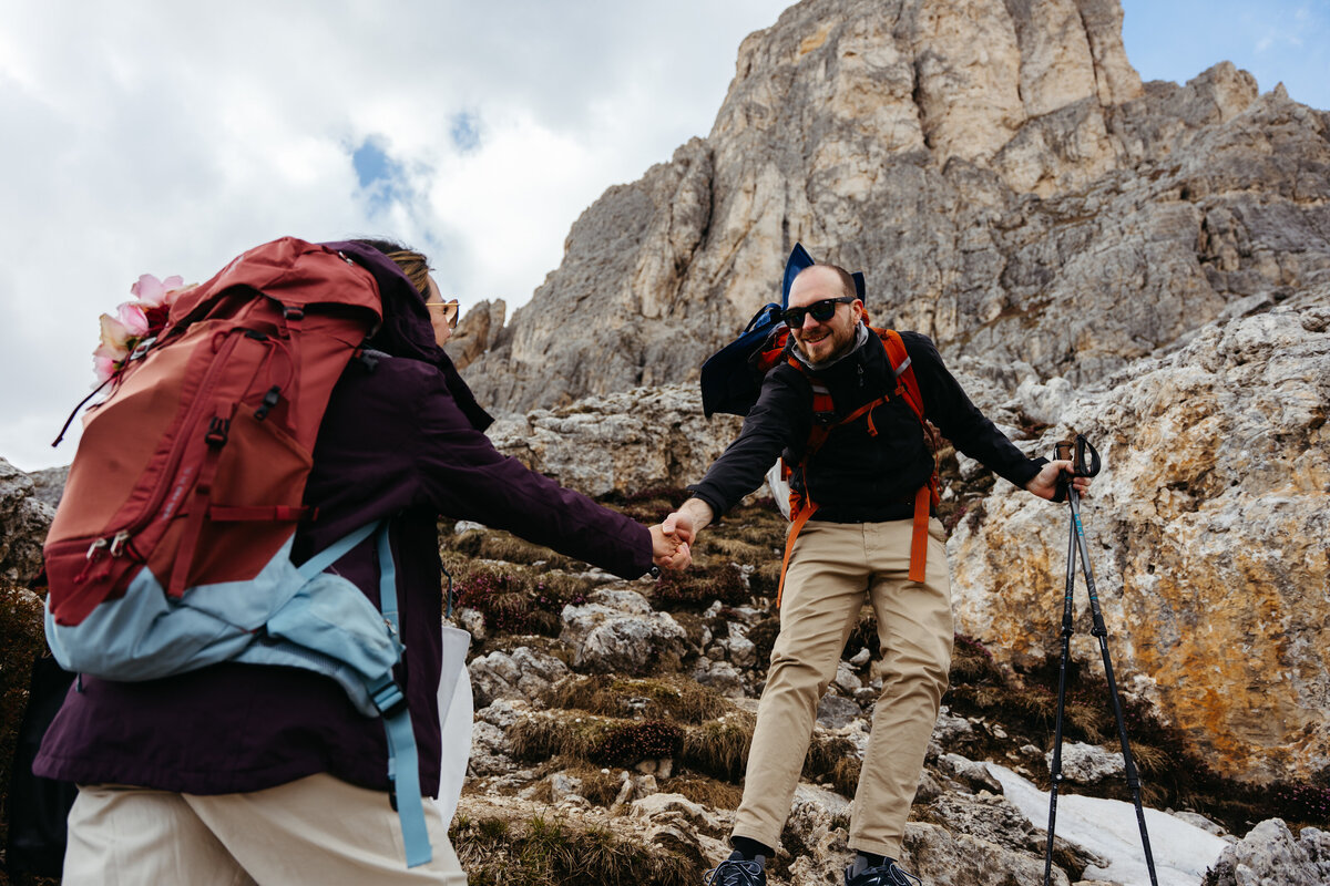 Groom helping bride climb rocky trail with backpacks