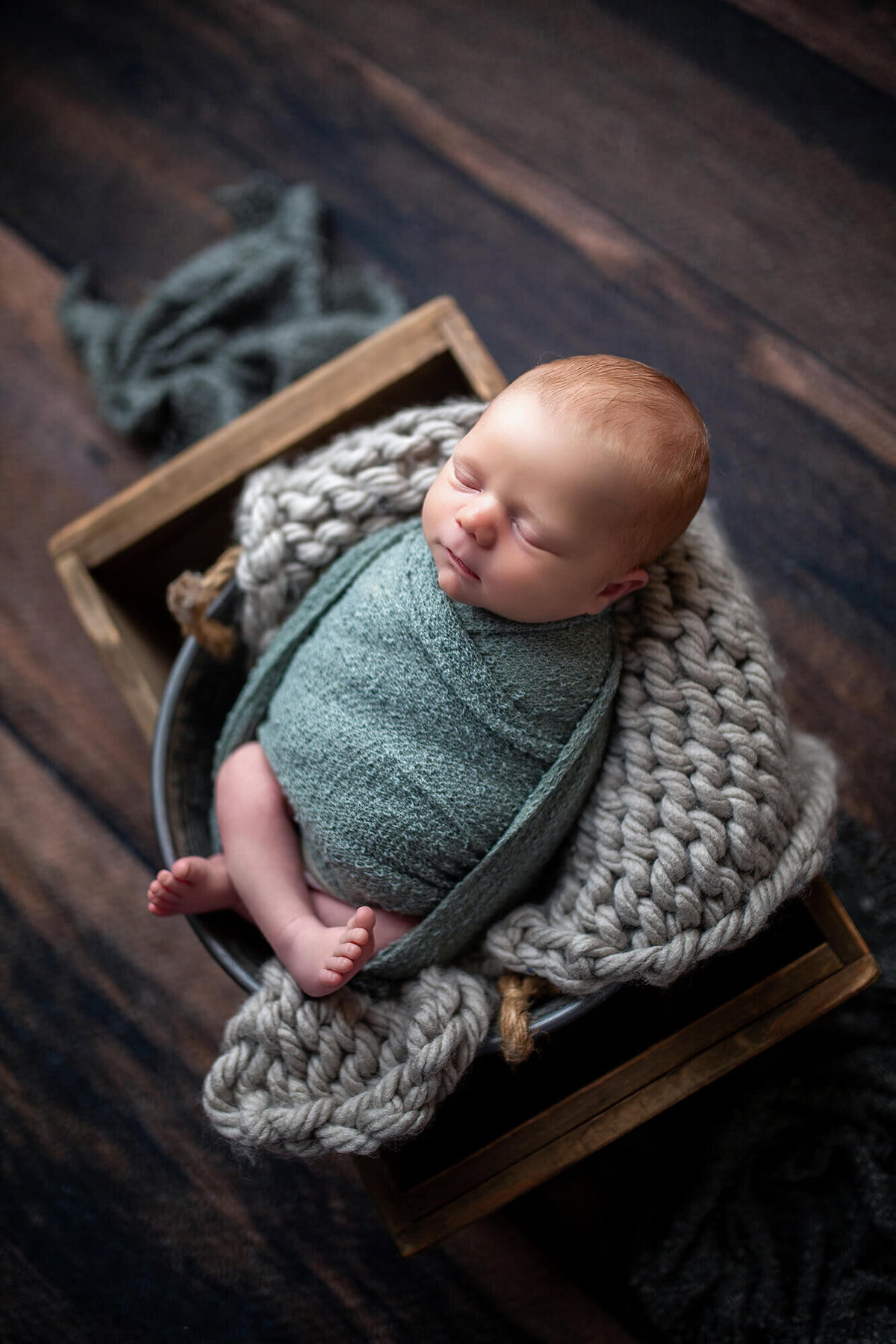 Baby boy wrapped in green with legs showing and sitting in grey bucket inside a crate
