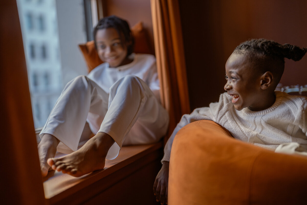 Modern and casual portrait of young brothers laughing and relaxing by a window, captured during a magazine-style photoshoot with a DFW family photographer. The boys are dressed in stylish, comfortable clothing, reflecting a relaxed and joyful moment in a contemporary setting.