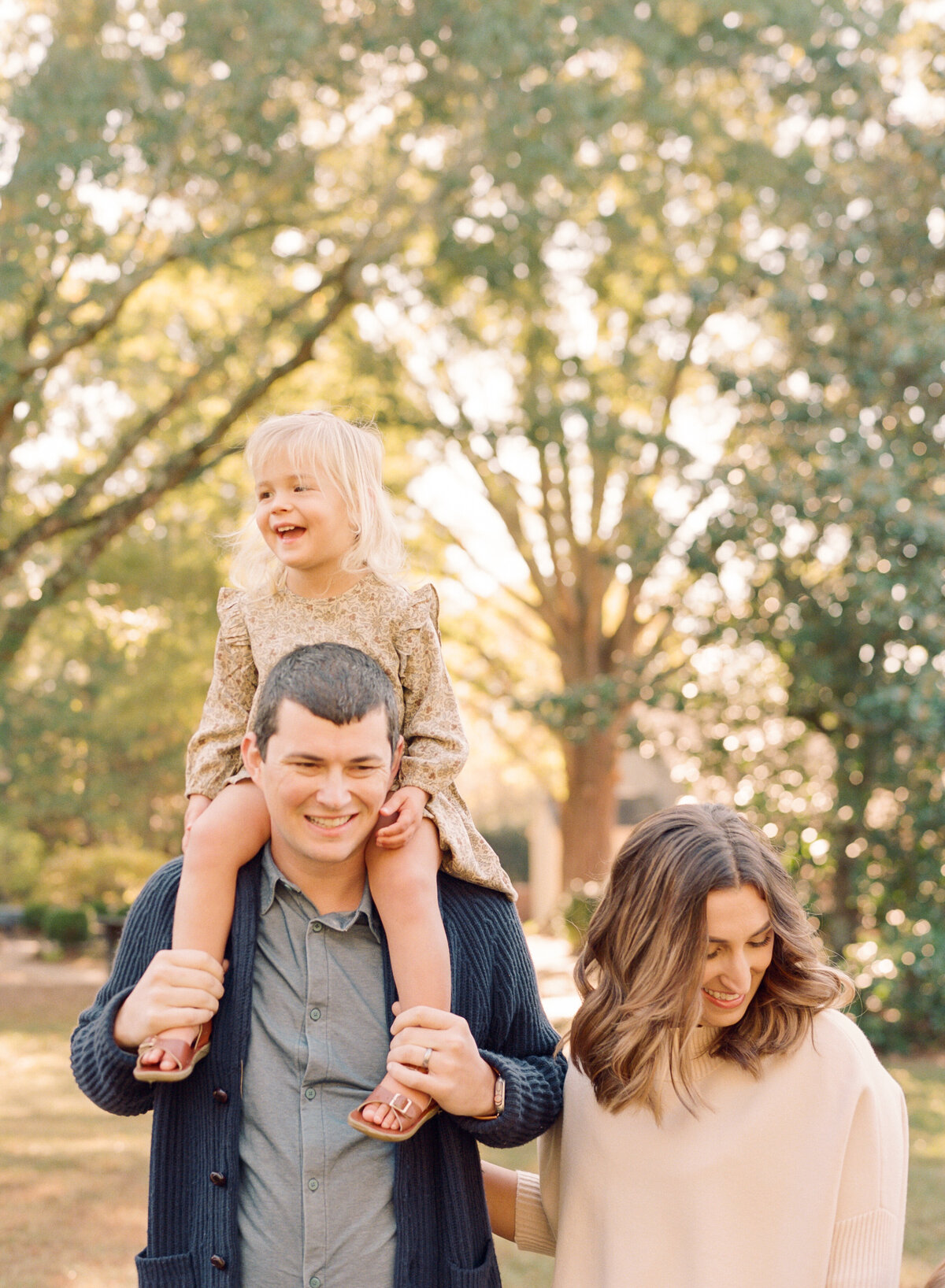 Toddler rides on her dad's shoulders during a fall family session in Raleigh. Family walking during their family portrait session in Wake Forest, NC. Photographed by Raleigh family photographer A.J. Dunlap Photography.