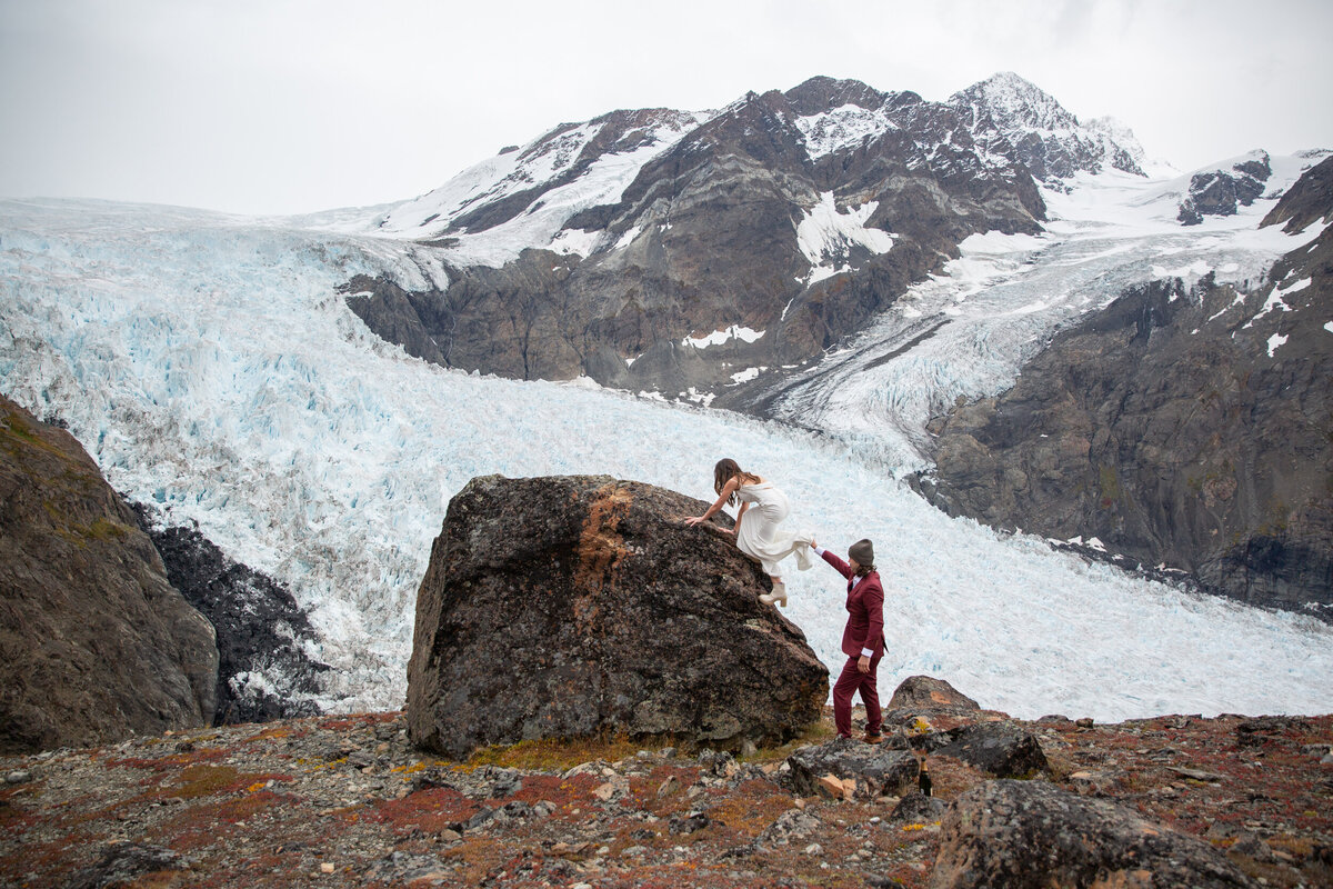 A groom helps his bride up a big boulder in Alaska with a snowy glacier behind them.