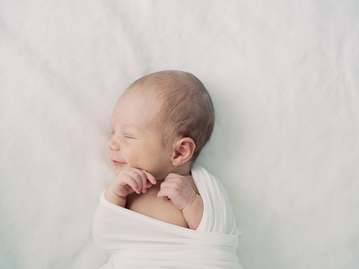 Newborn baby girl swaddled in white lays on a white bed and gives a soft newborn smile.