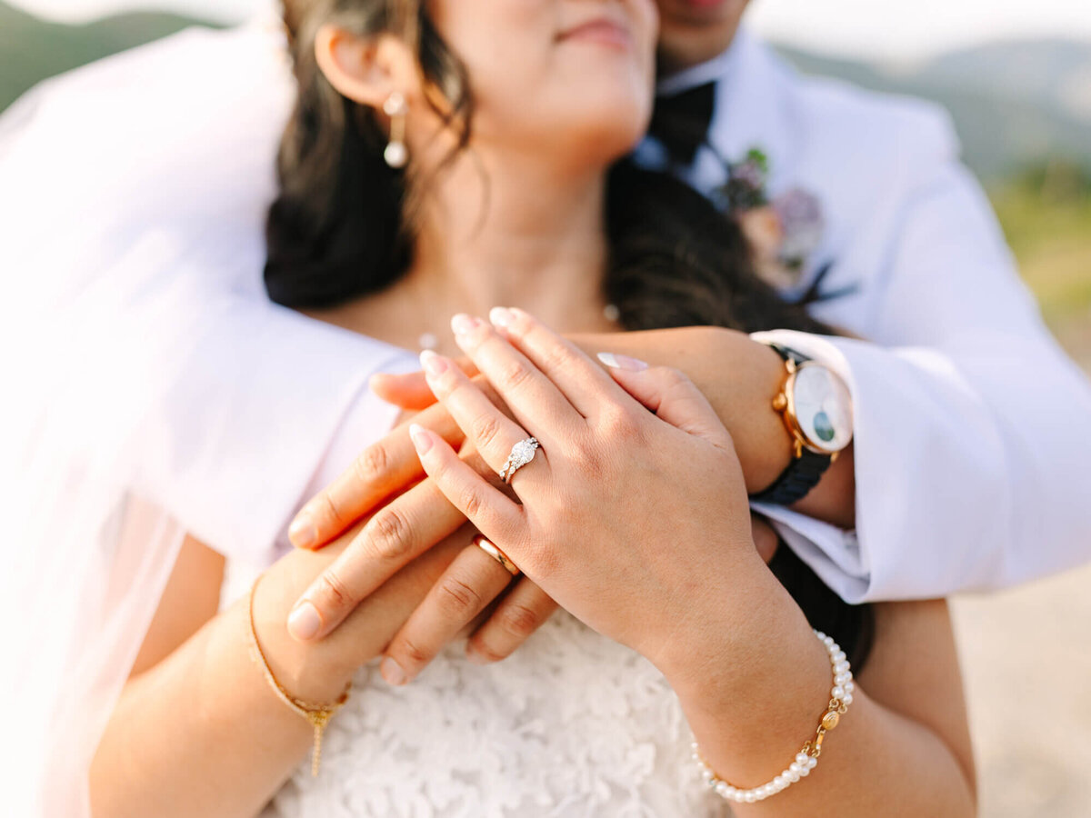 A bride and groom embrace, showcasing their hands adorned with rings and watches. The bride's lace dress and veil add elegance, conveying love and joy.
