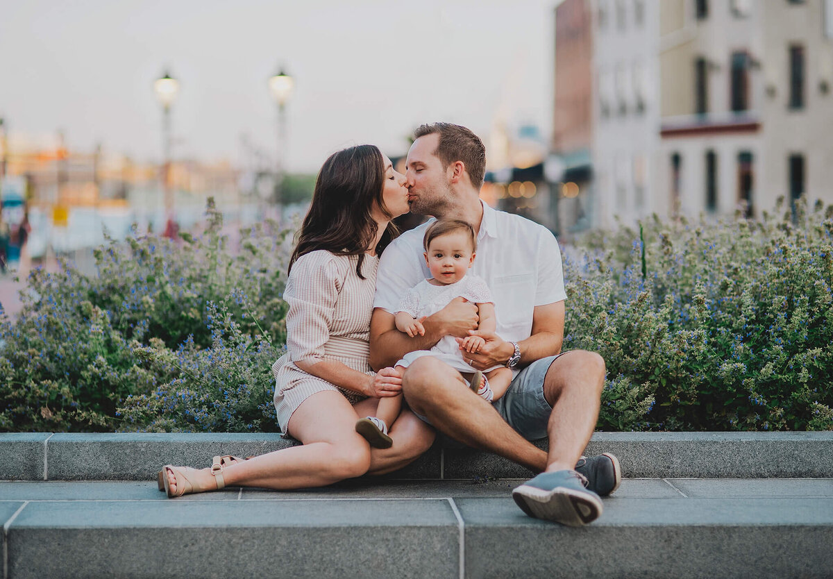 Mother and father kissing holding baby girl sitting in front of flowers in Fells Point Baltimore Maryland