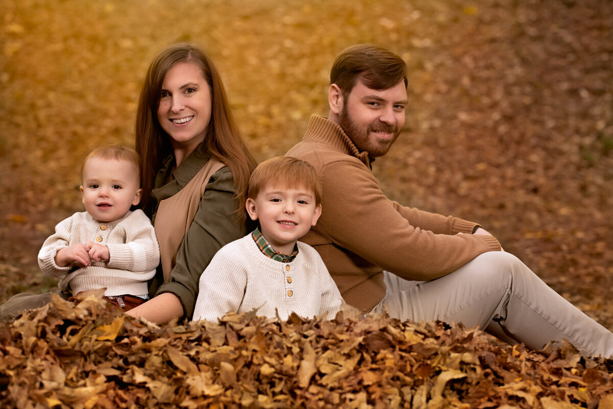 Warm autumn family portrait featuring parents and two children playing in leaves, captured in Overland Park, Kansas. Ideal for families seeking seasonal outdoor photography in the Kansas City metro area