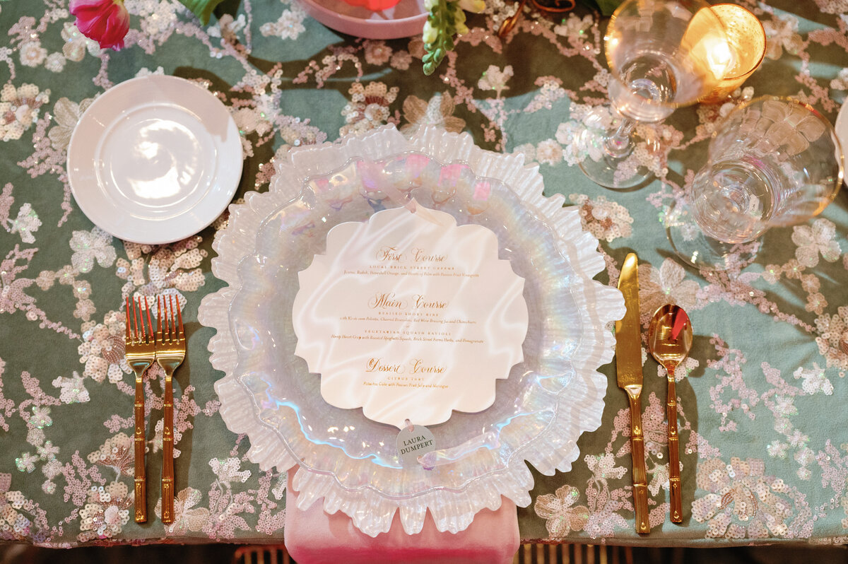 A refined close-up of a wedding menu card elegantly placed on a frosted white plate with coordinating white chargers and gold silverware. This sophisticated table setting blends timeless elegance with modern luxury, perfect for couples seeking a polished and stylish reception tablescape.