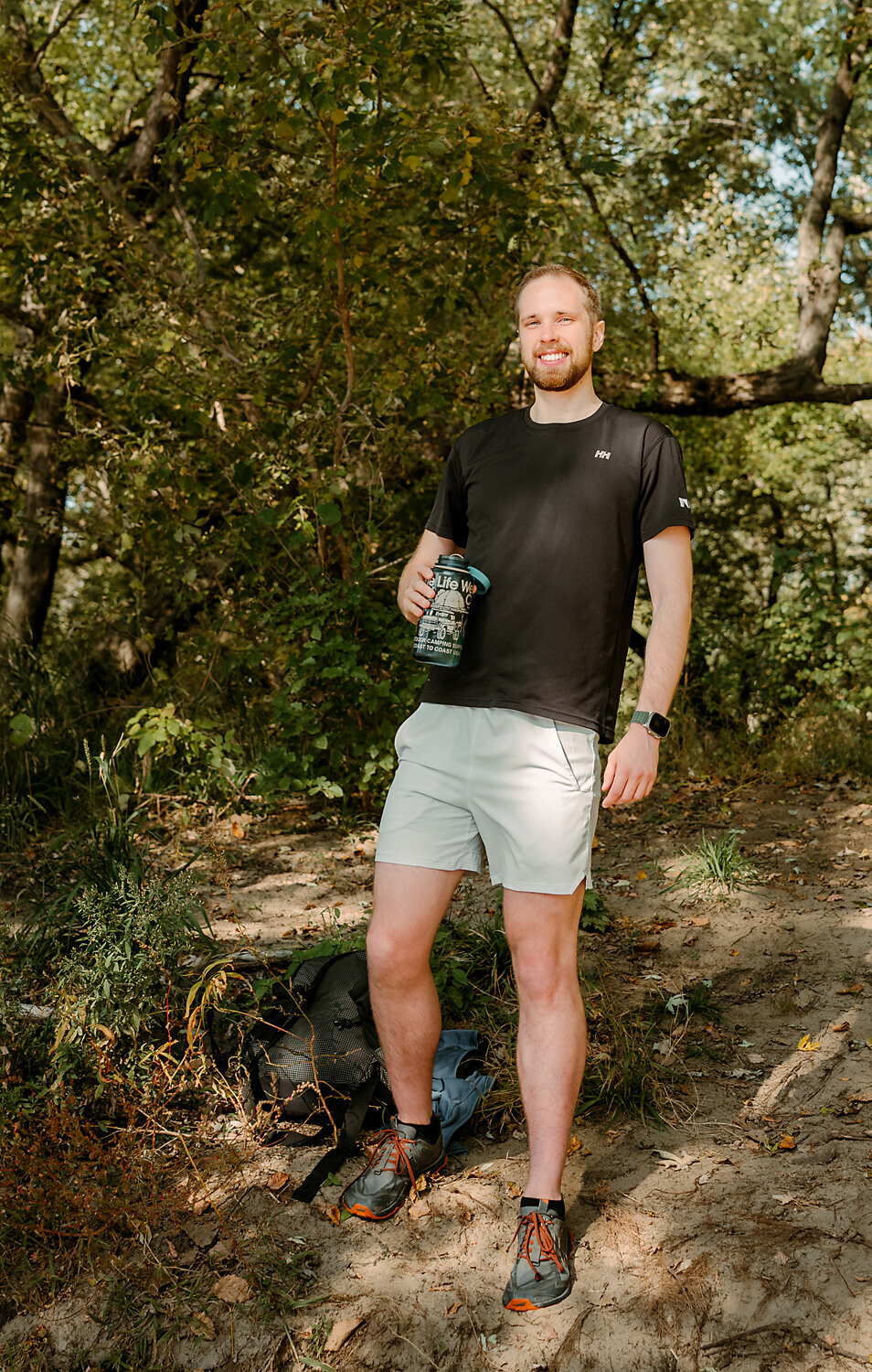 After photo of client standing confidently in a wooded outdoor setting, smiling and holding a water bottle during professional online dating photo session with Shannon Kathleen Photography.
