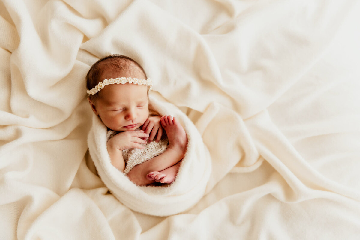 Baby wrapped in a white swaddle with hands under her chin while wearing a white floral headband.