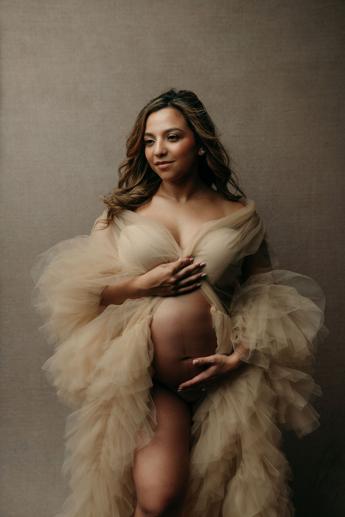 Pregnant mom wearing cream tulle dress open on belly holding baby bump on neutral backdrop