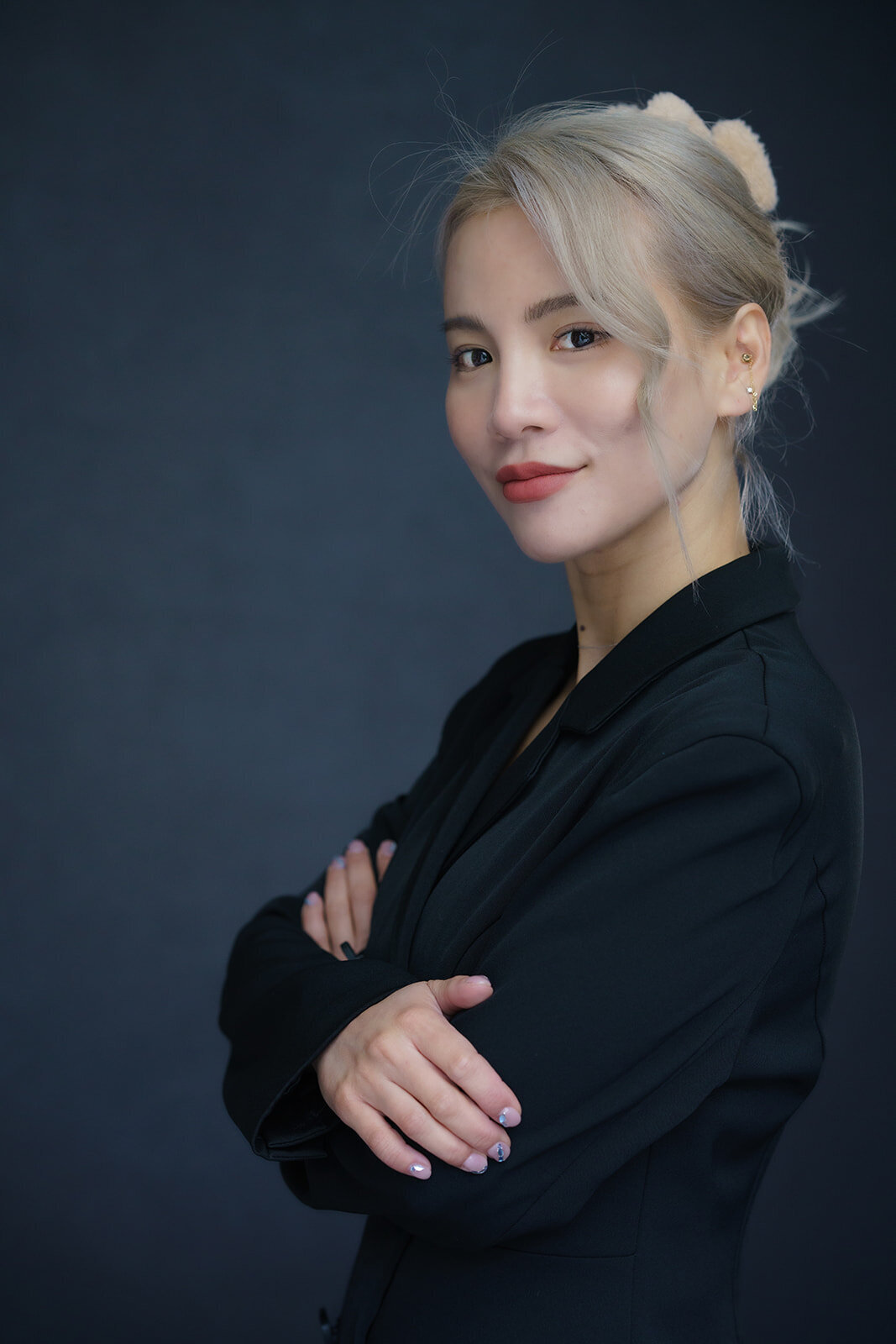 Side view of a Asian woman posed standing with arms crossed while wearing a dark suit and light colored hair 