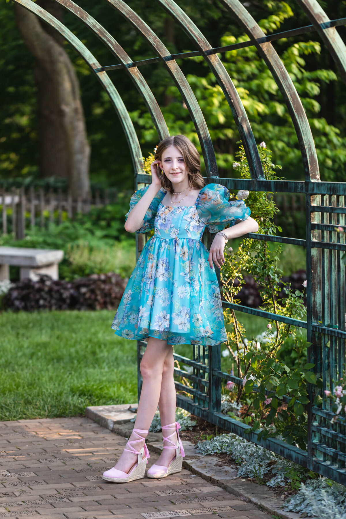 Lovely young teenage girl in a blue floral dress and pink shoes leaning on a garden trellis surrounded by roses. Captured by Springfield, MO teen photographer Dynae Levingston.
