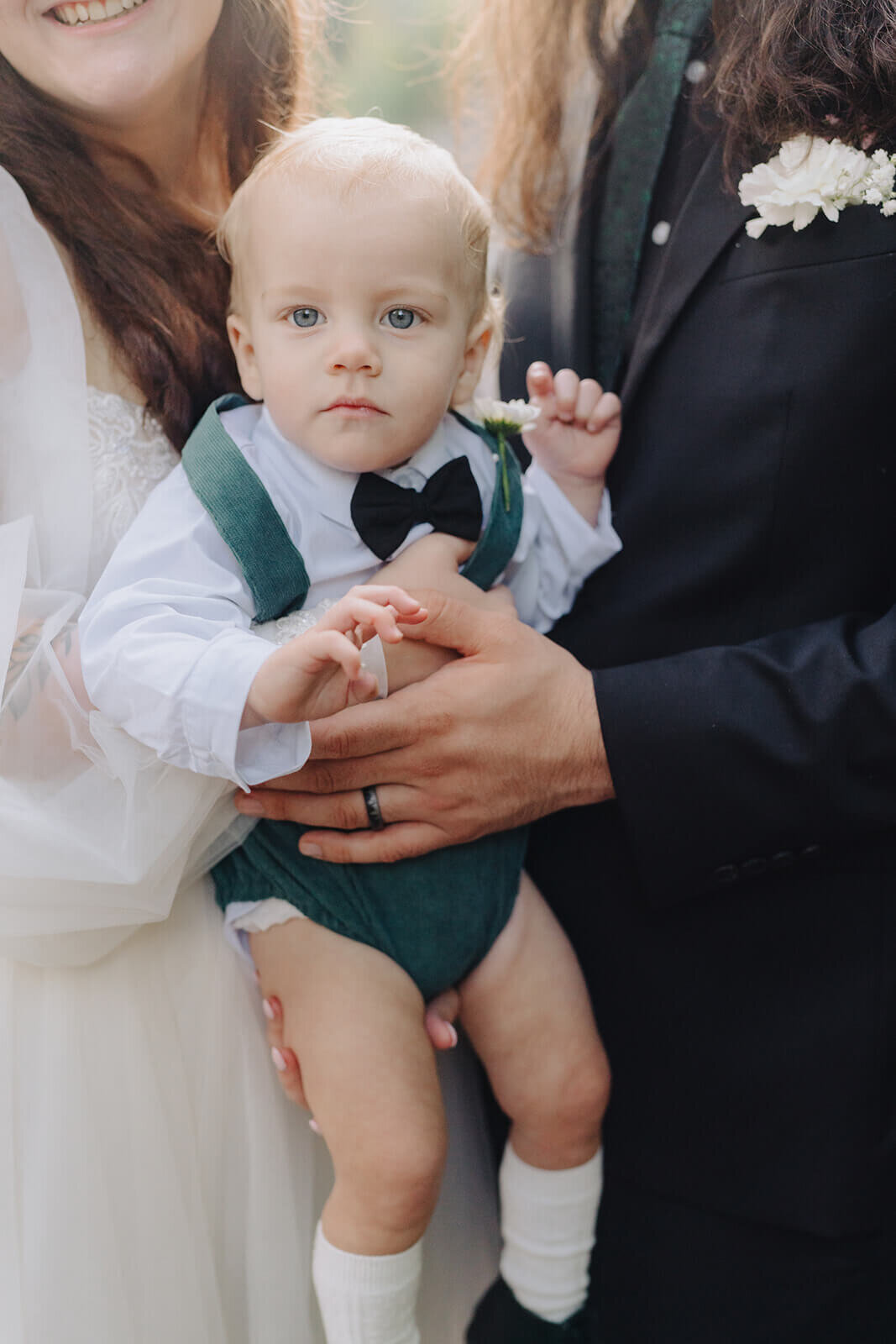 baby being held by bride and groom wearing bowtie, white shirt and green onesie