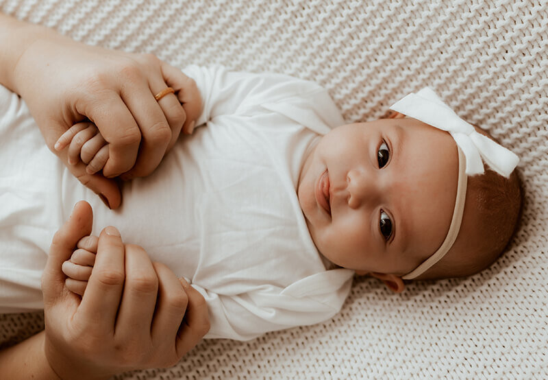Mom holding newborn baby girl's hands while laying on a white knit blanket wearing a white bow