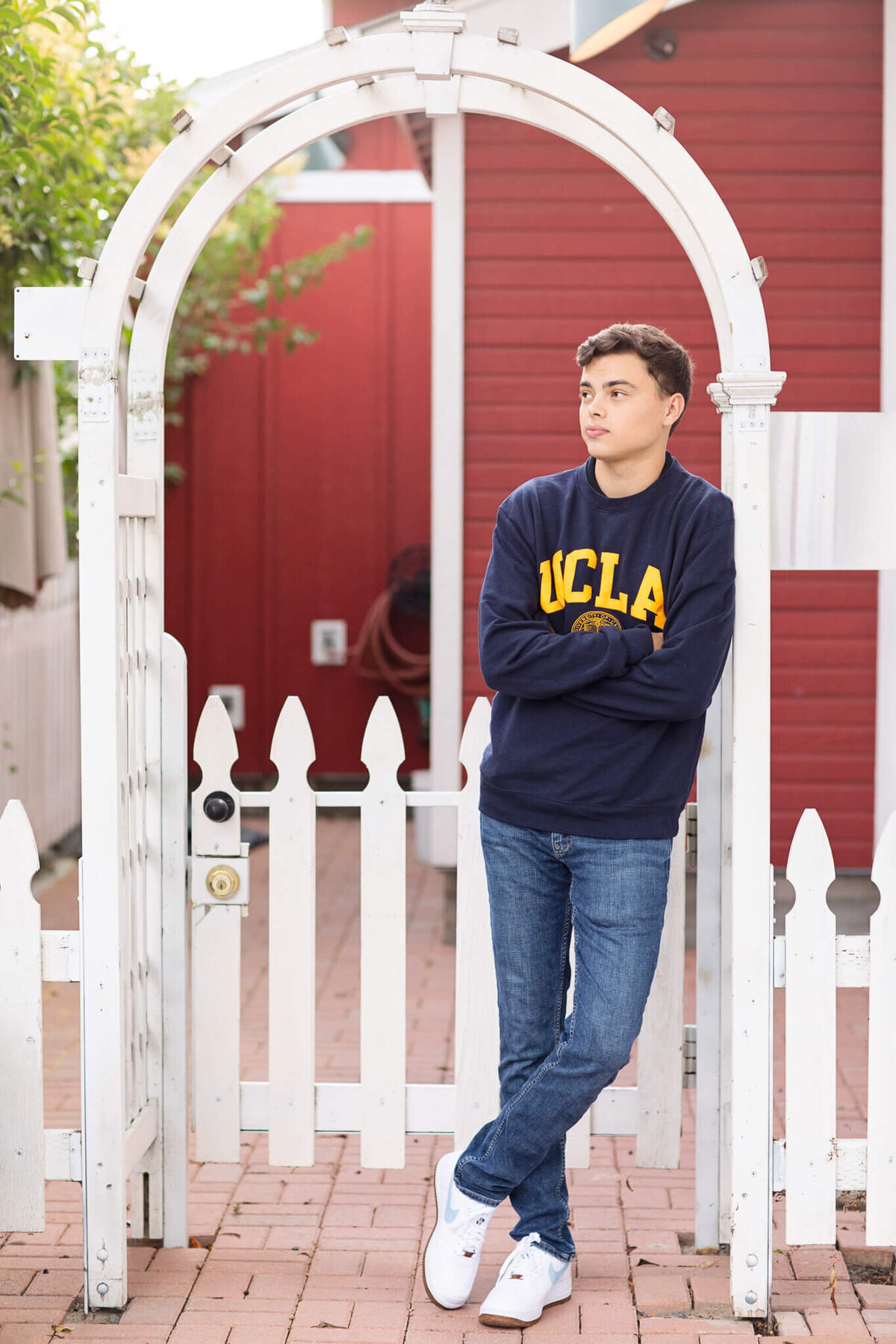 A person leans against a white picket gate, wearing a navy UCLA sweatshirt and jeans. They are gazing to the side, standing in front of a red building with a brick pathway underneath.