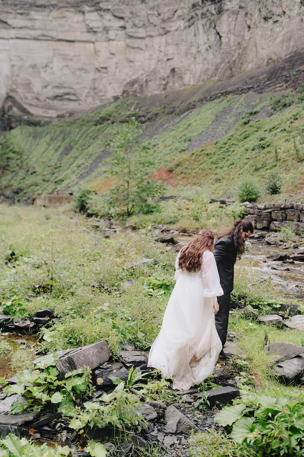 Bride and groom walking along rocks and greenery at Finger Lakes wedding, NY