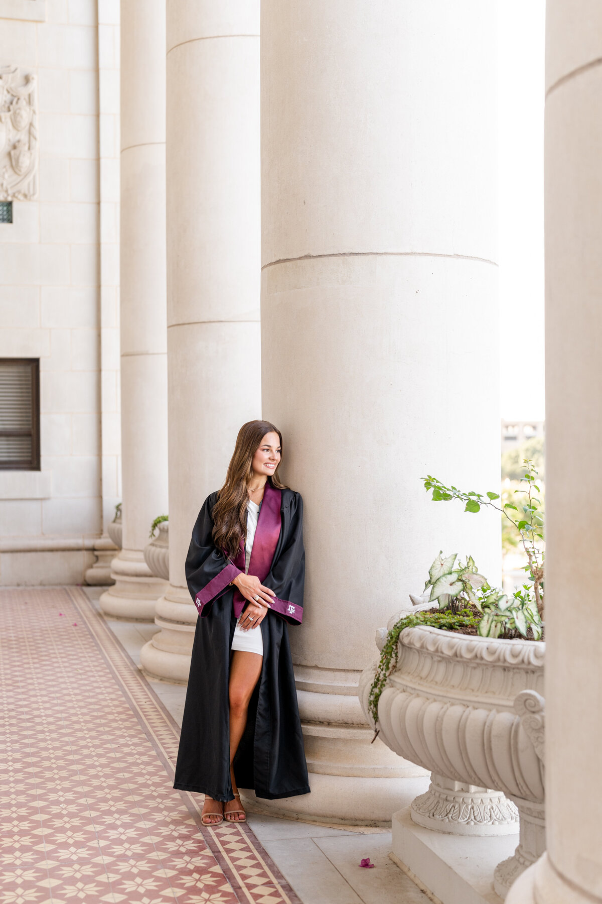 Texas A&M senior girl wearing gown and Aggie stole while holding wrist and leaning against column and looking away at Administration Building
