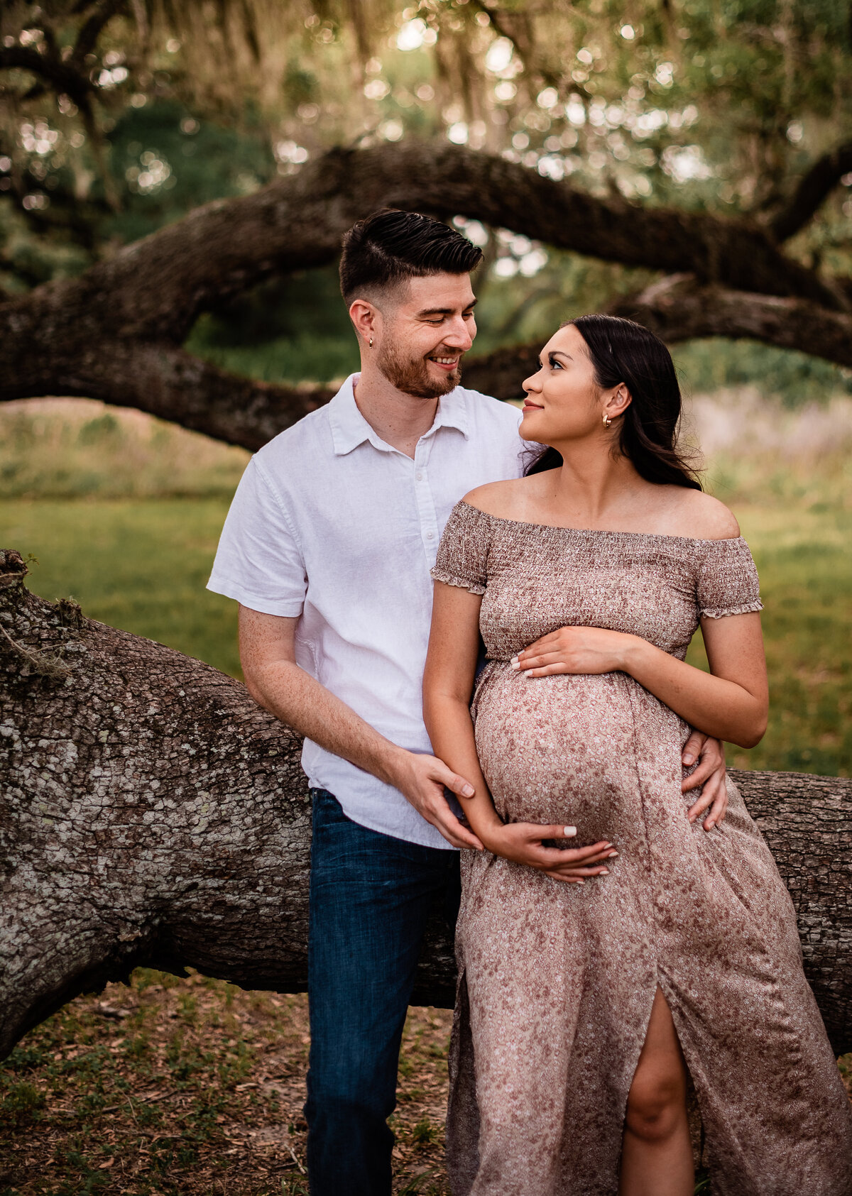 A pregnant couple leans on a low oak tree branch while looking at each other.