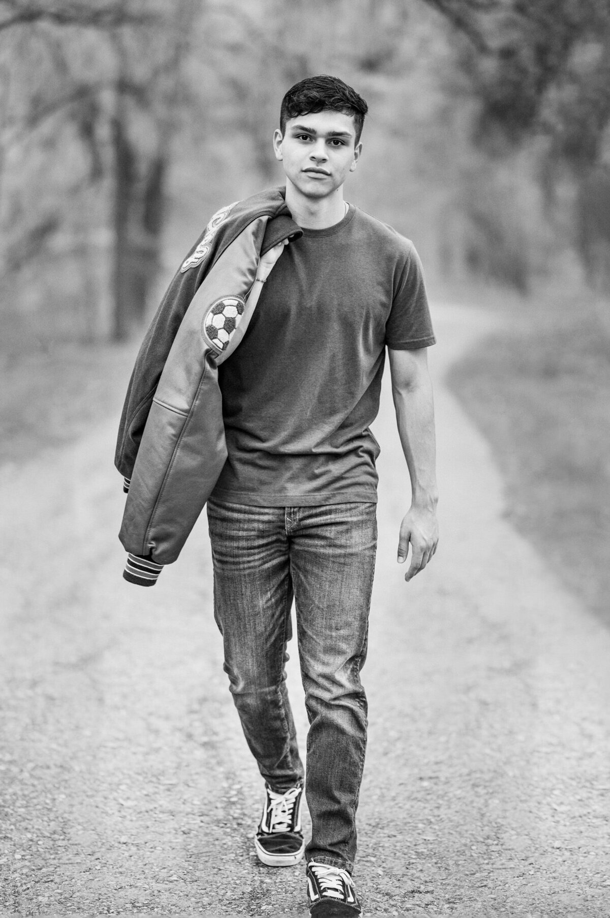 Black and white image of a San Antonio senior. His letter jacket is thrown over his shoulder and he's walking towards the camera.