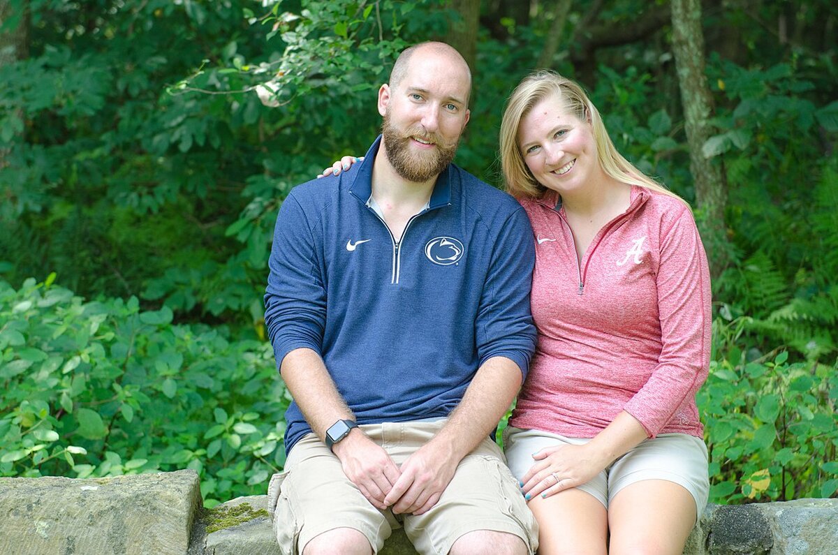 An Alabama Crimson Tide and Penn State fan engaged couple sit on a rock wall at Twin Lakes Park in Greensburg, PA