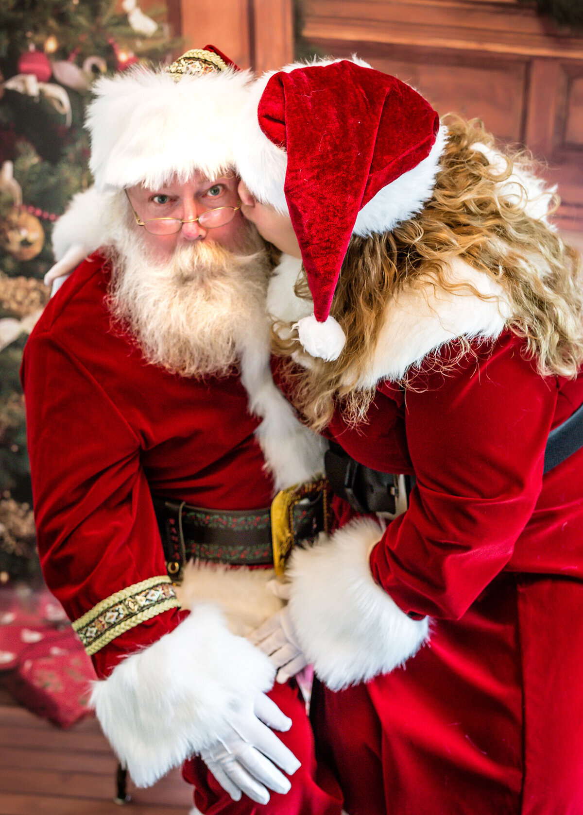 Mrs. Claus kisses Santa Claus on the cheek & he looks surprised while posing for a South Shore MA branding photographer